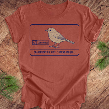 A rust-orange crewneck t-shirt featuring a graphic of a little brown bird with a 'Confirmed' stamp and the text 'CLASSIFICATION: LITTLE BROWN JOB (LB)'. The shirt is laid flat on a rustic wood background with pine needles in the corners.