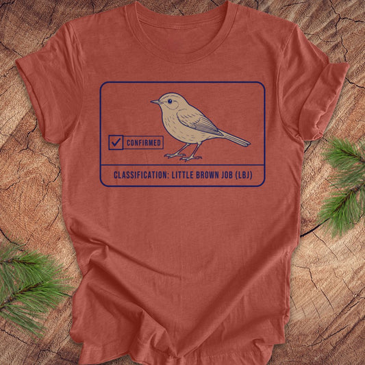 A rust-orange crewneck t-shirt featuring a graphic of a little brown bird with a 'Confirmed' stamp and the text 'CLASSIFICATION: LITTLE BROWN JOB (LB)'. The shirt is laid flat on a rustic wood background with pine needles in the corners.