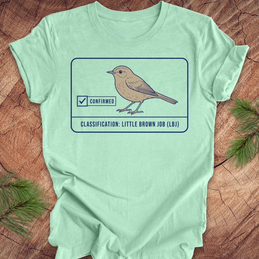 A mint green short-sleeve t-shirt laid flat on a rustic wood surface with pine branches in the corners. The shirt features a graphic of a little brown job bird with a 'Confirmed' checkmark and the text 'CLASSIFICATION: LITTLE BROWN JOB (LBJ)'.