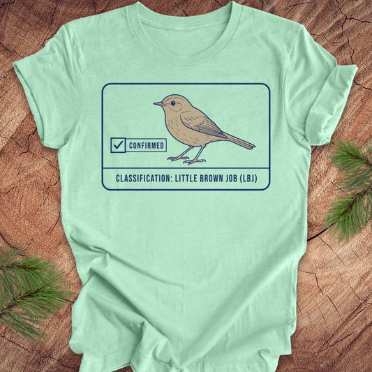 A mint green short-sleeve t-shirt laid flat on a rustic wood surface with pine branches in the corners. The shirt features a graphic of a little brown job bird with a 'Confirmed' checkmark and the text 'CLASSIFICATION: LITTLE BROWN JOB (LBJ)'.