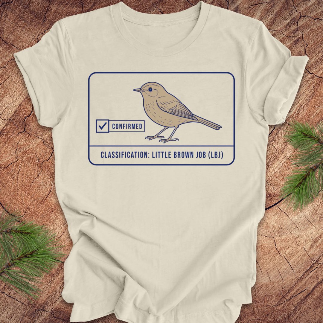 An beige short-sleeve t-shirt laid flat on a rustic wood background with pine branches in the corners. The shirt features a graphic of a little brown job bird with a 'Confirmed' checkmark and the text 'CLASSIFICATION: LITTLE BROWN JOB (LBI)'.