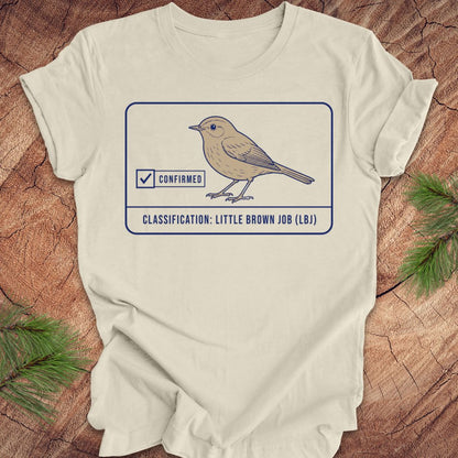 An beige short-sleeve t-shirt laid flat on a rustic wood background with pine branches in the corners. The shirt features a graphic of a little brown job bird with a 'Confirmed' checkmark and the text 'CLASSIFICATION: LITTLE BROWN JOB (LBI)'.
