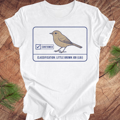 A white short-sleeve T-shirt laid flat on a rustic wood surface with pine branches at the corners. The shirt has a printed graphic of a small brown bird inside a bordered box with the words 'CONFIRMED' and 'CLASSIFICATION: LITTLE BROWN JOB (LB)'.