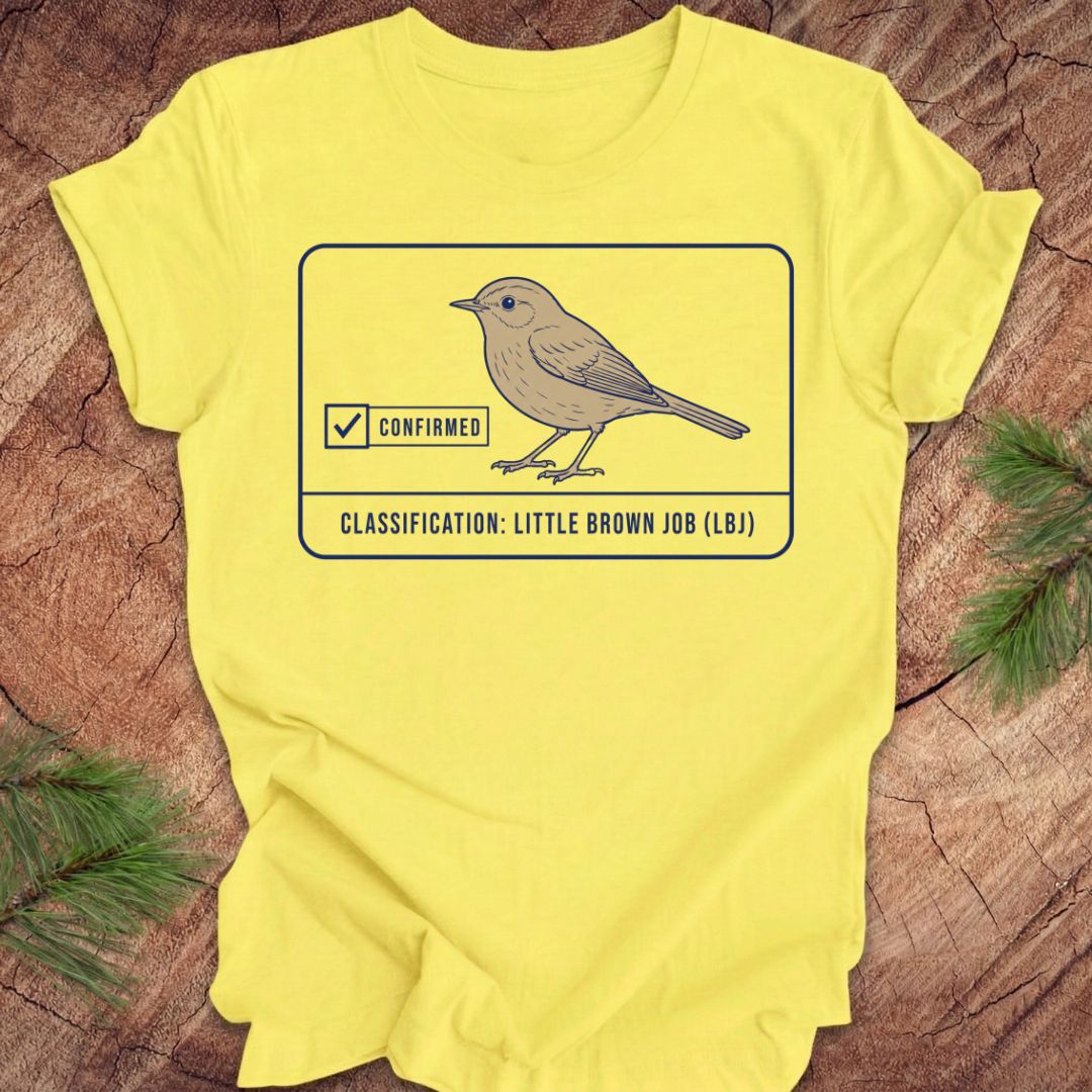 A bright yellow short-sleeve t-shirt laid flat on a rustic wood surface with pine branches at the corners. The front features a graphic of a little brown bird inside a bordered box with a checked box and the text 'CLASSIFICATION: LITTLE BROWN JOB (LB)'.