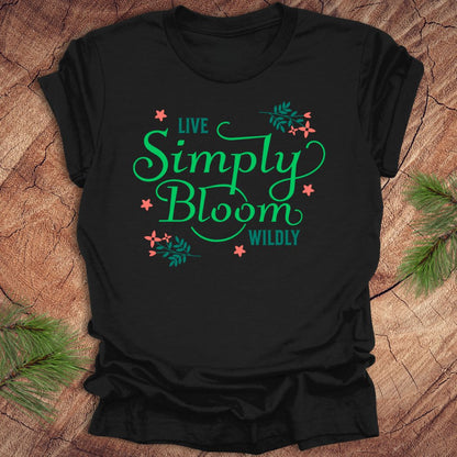 Black t-shirt with 'Live Simply Bloom Wildly' text on a wooden surface with greenery.