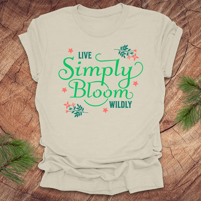 Beige t-shirt with 'Live Simply Bloom Wildly' text on a wooden surface with greenery.