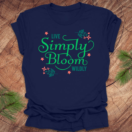 Navy blue t-shirt with 'Live Simply Bloom Wildly' text on a wooden surface with greenery.