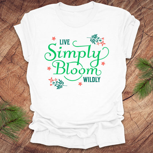 White t-shirt with 'Live Simply Bloom Wildly' text on a wooden surface with greenery.