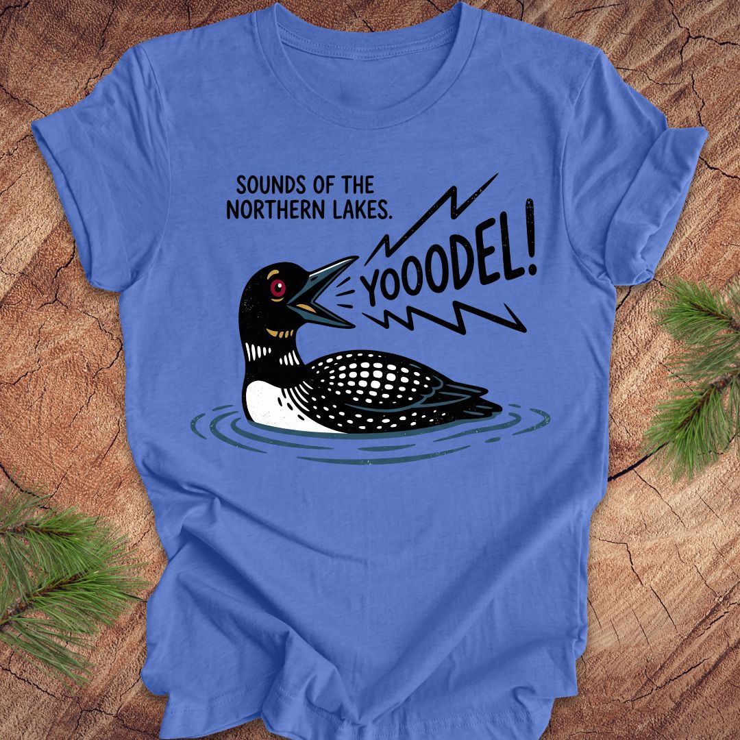 A blue short-sleeve crewneck t-shirt featuring a graphic of a loon with the text 'SOUNDS OF THE NORTHERN LAKES. YOOODEL!' in black, shown laid flat on a rustic wood background with pine needles.