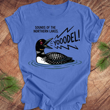 A blue short-sleeve crewneck t-shirt featuring a graphic of a loon with the text 'SOUNDS OF THE NORTHERN LAKES. YOOODEL!' in black, shown laid flat on a rustic wood background with pine needles.