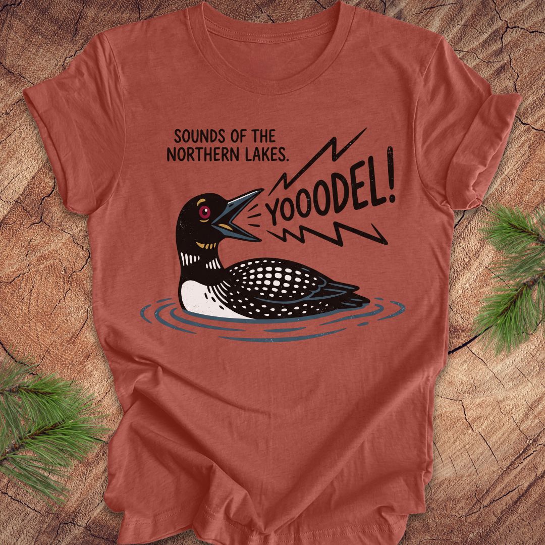 A terracotta-colored short-sleeve t-shirt with a graphic of a loon and the text 'SOUNDS OF THE NORTHERN LAKES. YOOODEL!' laid flat on a rustic wood background with pine branches.