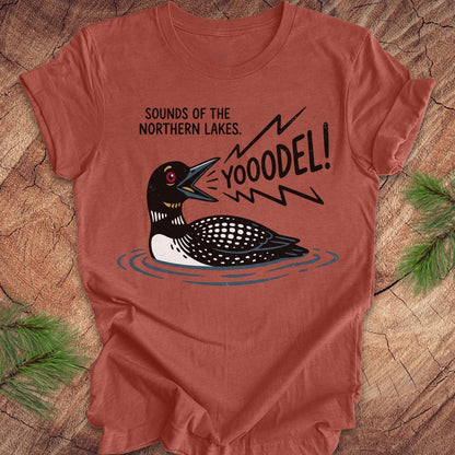 A terracotta-colored short-sleeve t-shirt with a graphic of a loon and the text 'SOUNDS OF THE NORTHERN LAKES. YOOODEL!' laid flat on a rustic wood background with pine branches.