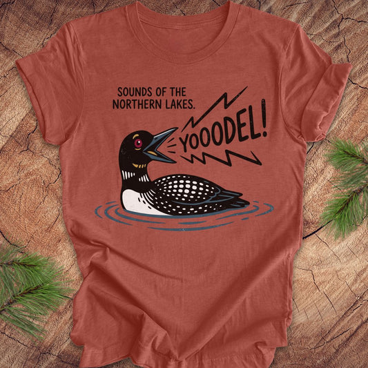 A terracotta-colored short-sleeve t-shirt with a graphic of a loon and the text 'SOUNDS OF THE NORTHERN LAKES. YOOODEL!' laid flat on a rustic wood background with pine branches.
