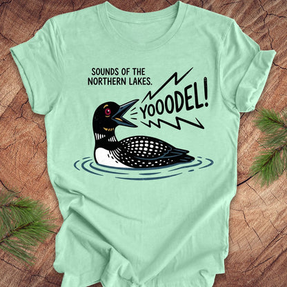 A mint green graphic t-shirt laid flat on a rustic wood background with pine needles in the corners. The shirt features a cartoon loon with the text 'SOUNDS OF THE NORTHERN LAKES.