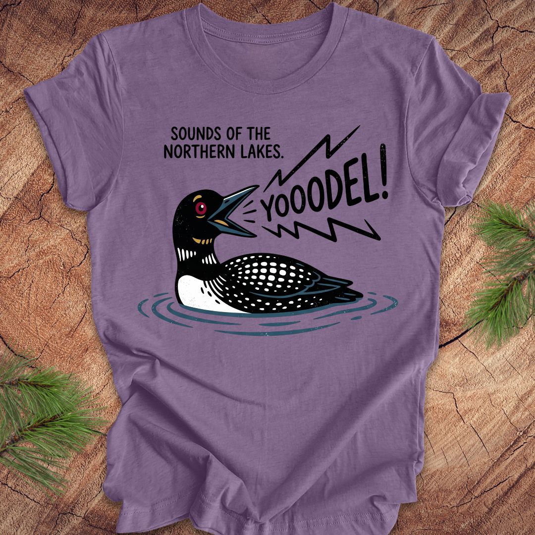 A purple short-sleeve t-shirt laid flat on a rustic wood background with pine branches in the corners. The shirt features a graphic of a loon with the text 'SOUNDS OF THE NORTHERN LAKES.