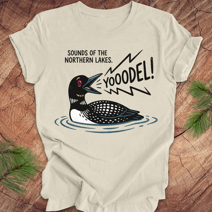 A cream-colored short-sleeve t-shirt with a graphic of a loon and the text 'SOUNDS OF THE NORTHERN LAKES. YOODEL!' printed on the front, displayed on a rustic wood background with pine branches.