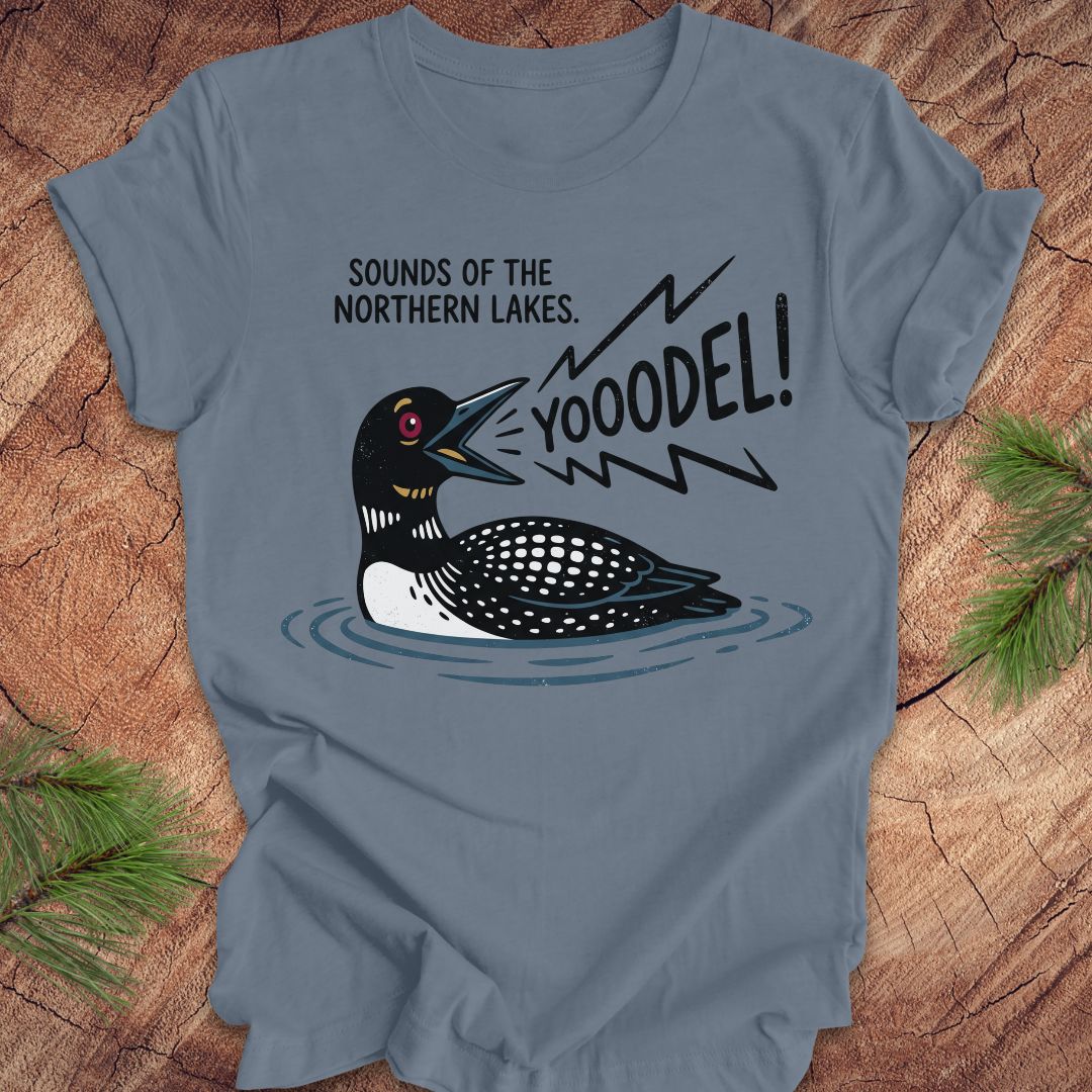 A slate blue crewneck t-shirt featuring a cartoon loon graphic with the text 'SOUNDS OF THE NORTHERN LAKES. YOOODEL!' on the front, displayed flat on a rustic wood background with pine needles.