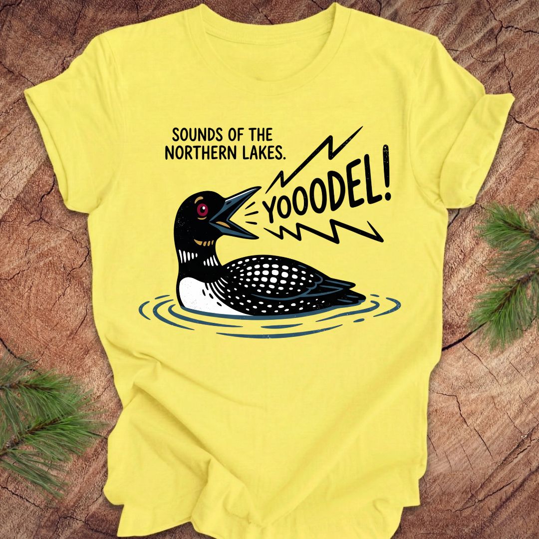 A bright yellow short-sleeve t-shirt laid flat on a rustic wood surface with pine branches at the corners. The shirt features a graphic of a loon in water with the text 'SOUNDS OF THE NORTHERN LAKES.