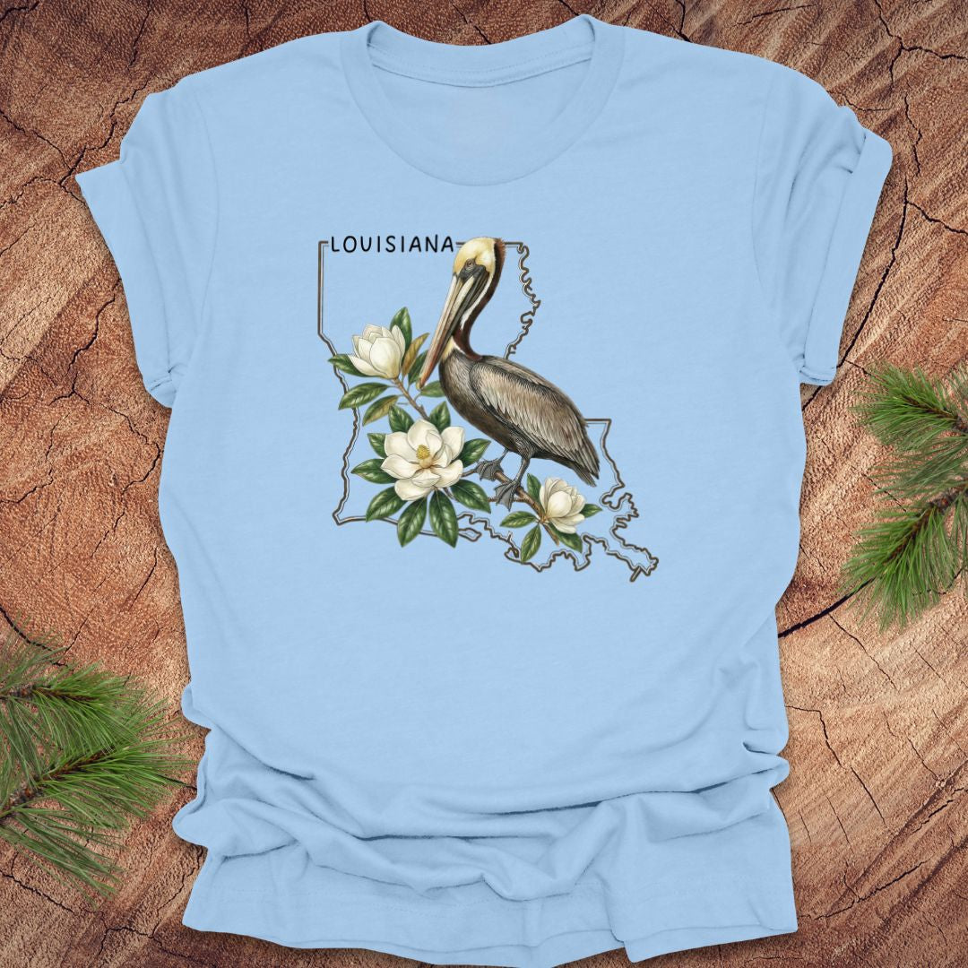 Light blue t-shirt with a Louisiana-themed design featuring a pelican and magnolias on a wooden surface.