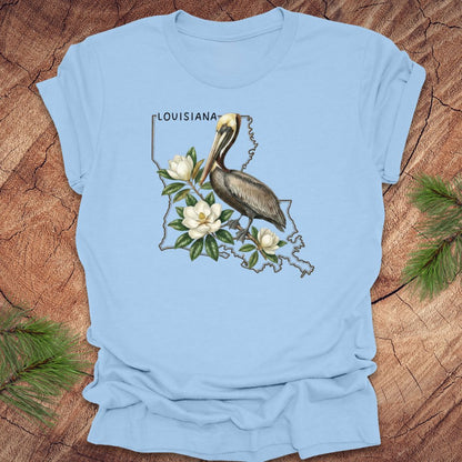 Light blue t-shirt with a Louisiana-themed design featuring a pelican and magnolias on a wooden surface.