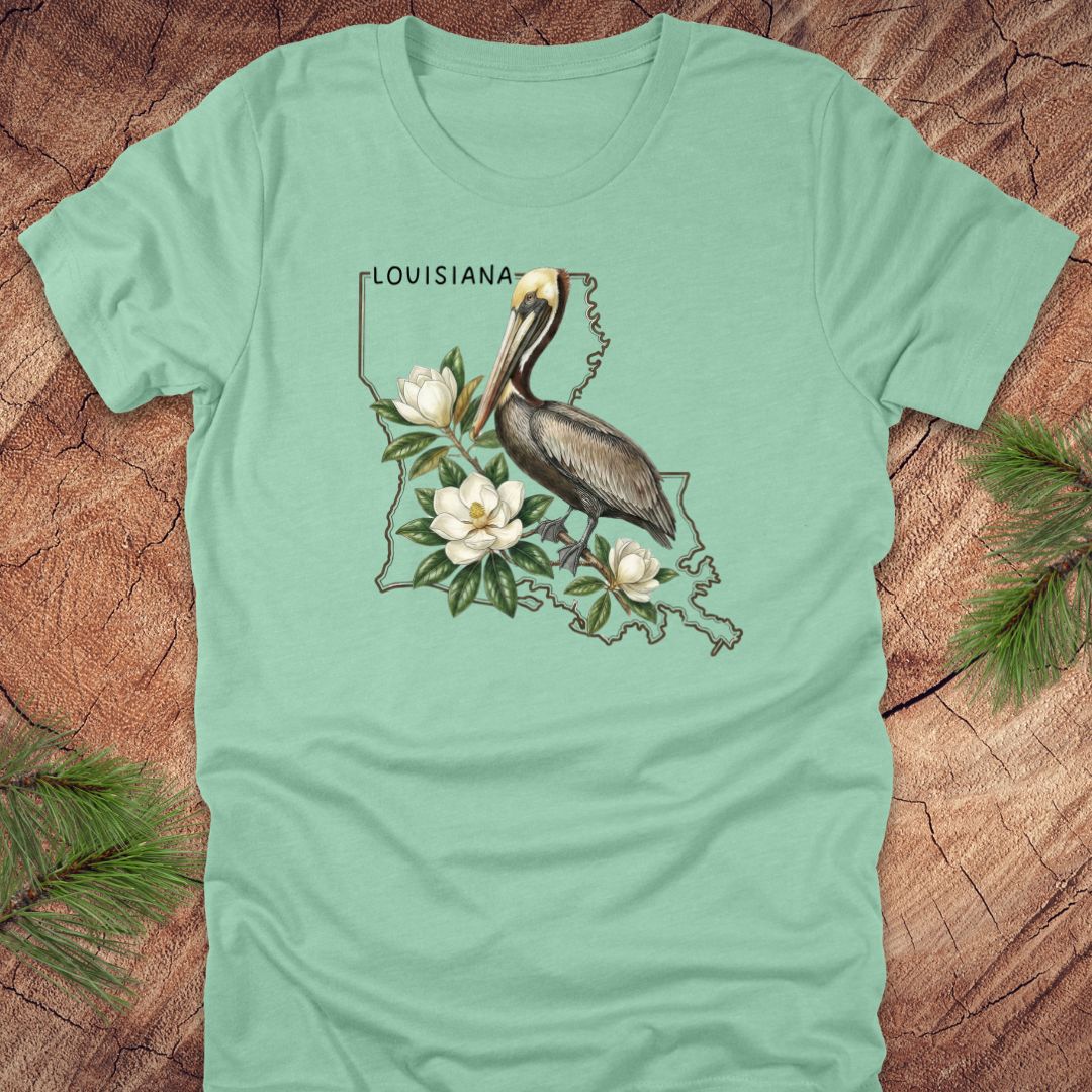Light green t-shirt with Louisiana-themed design featuring a pelican and magnolias on a wooden surface.