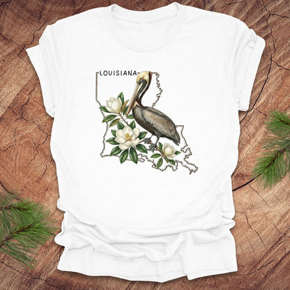White t-shirt with Louisiana-themed design featuring a pelican and magnolias on a wooden surface.
