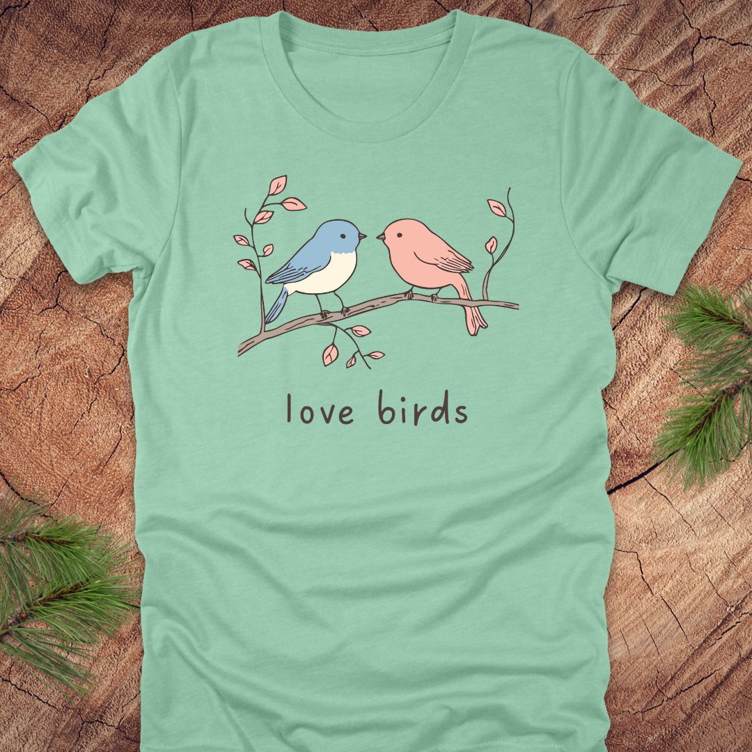 Mint green t-shirt with a blue and a pink hand drawn bird, on a branch, with text 'love birds' on a wooden background.