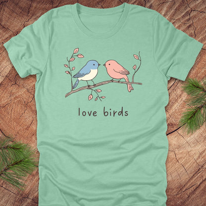 Mint green t-shirt with a blue and a pink hand drawn bird, on a branch, with text 'love birds' on a wooden background.