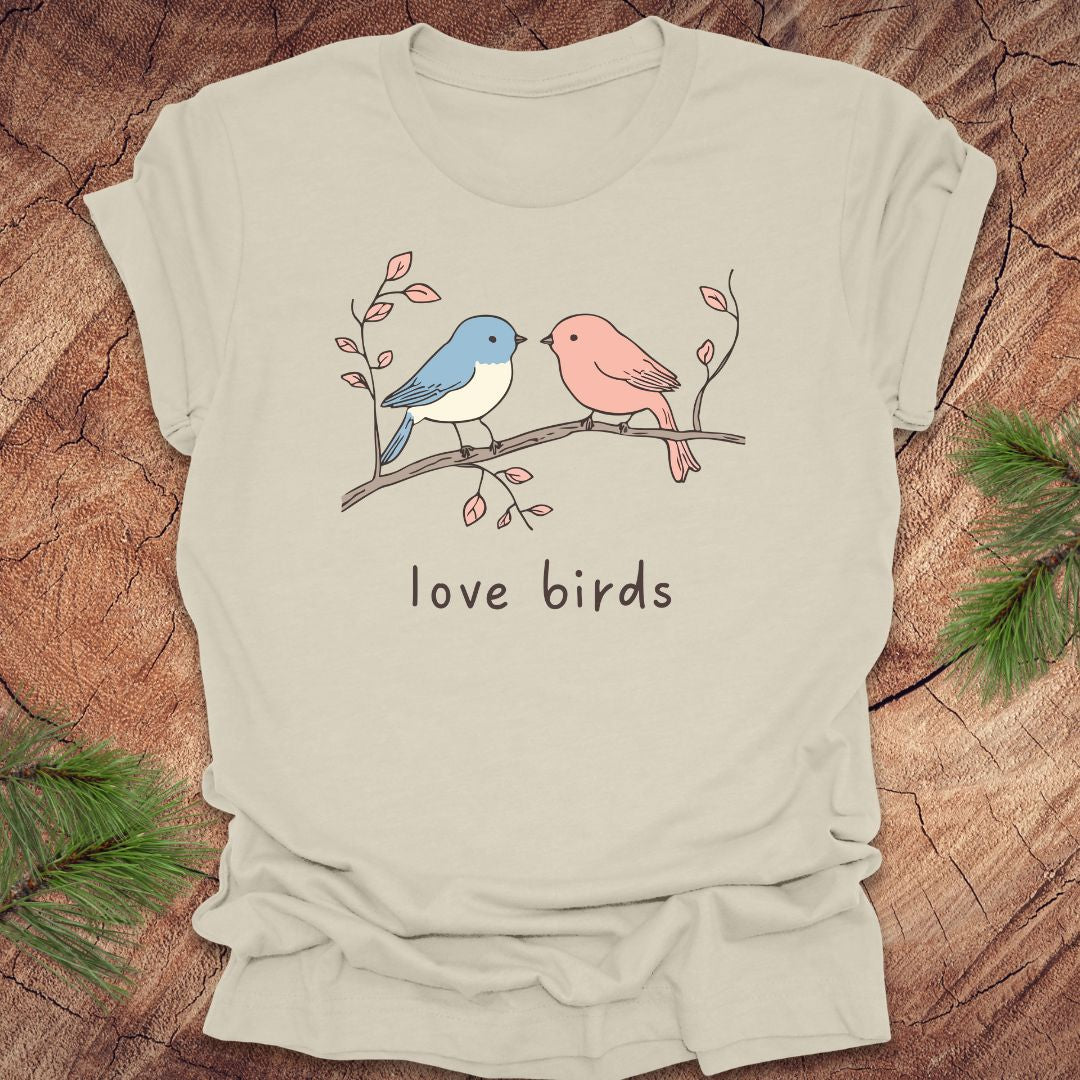 Beige t-shirt with a blue and a pink hand drawn bird, on a branch, with text 'love birds' on a wooden background.