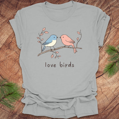 Silver t-shirt with a blue and a pink hand drawn bird, on a branch, with text 'love birds' on a wooden background.