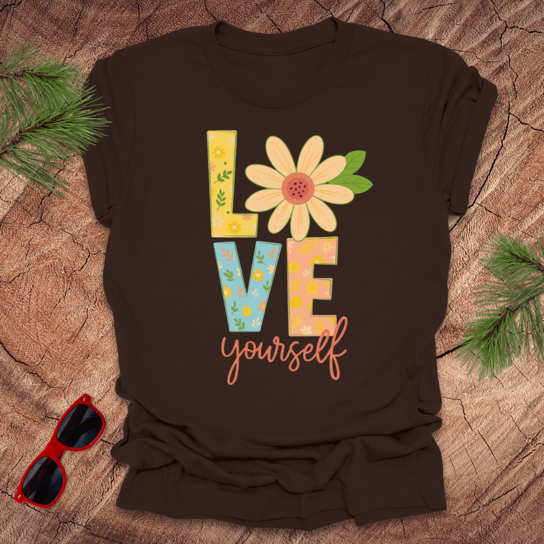Brown t-shirt with 'LOVE yourself' design on a wooden surface with sunglasses and greenery.