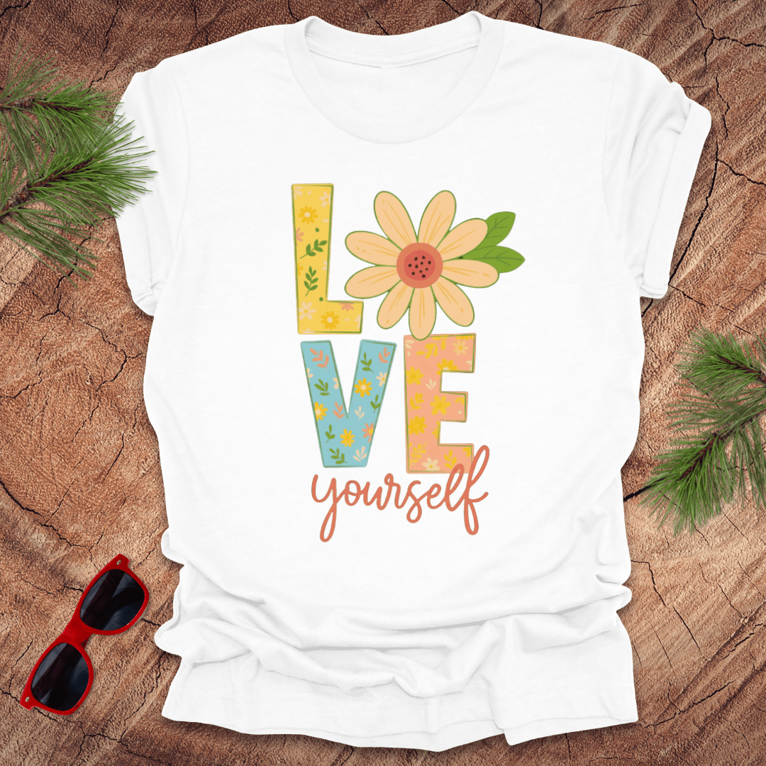 White t-shirt with 'Love yourself' design on a wooden surface with sunglasses and greenery.