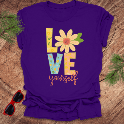 Purple t-shirt with 'Love yourself' text and flower design on a wooden surface with sunglasses and greenery.