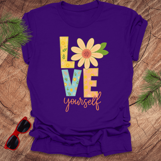 Purple t-shirt with 'Love yourself' text and flower design on a wooden surface with sunglasses and greenery.