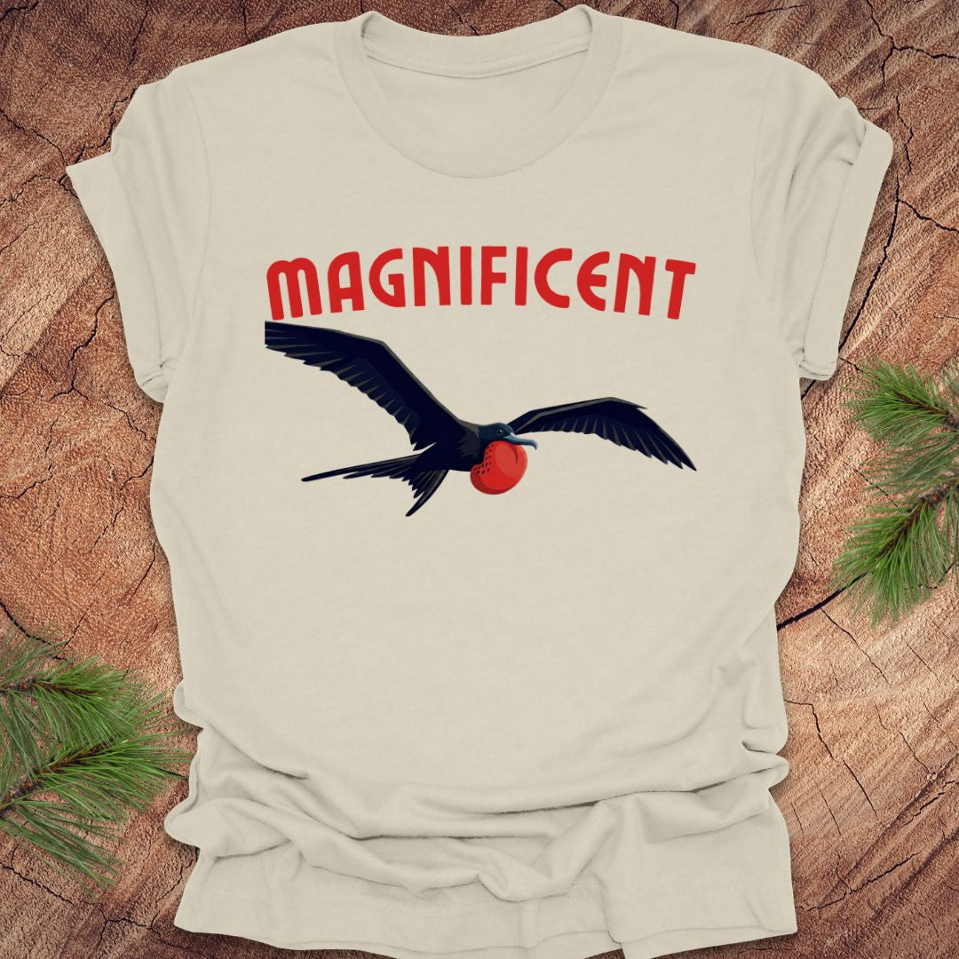 Beige t-shirt with a bird graphic and 'MAGNIFICENT' text on a wooden surface.