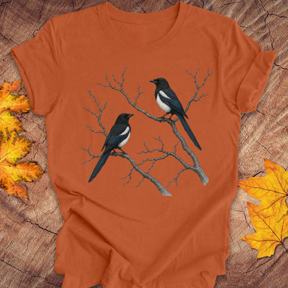 An orange short-sleeve t-shirt with a graphic of two magpies perched on bare branches, laid flat on a rustic wood background with autumn leaves.