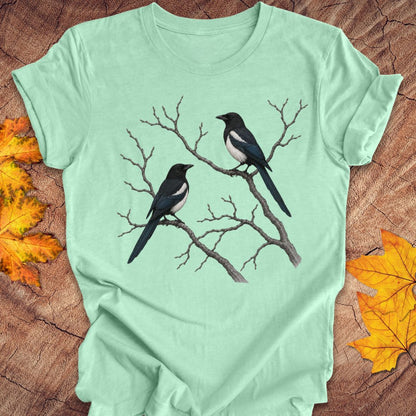 A mint green short-sleeve t-shirt featuring a graphic of two magpies on bare tree branches, laid flat on a rustic wood background with autumn leaves.