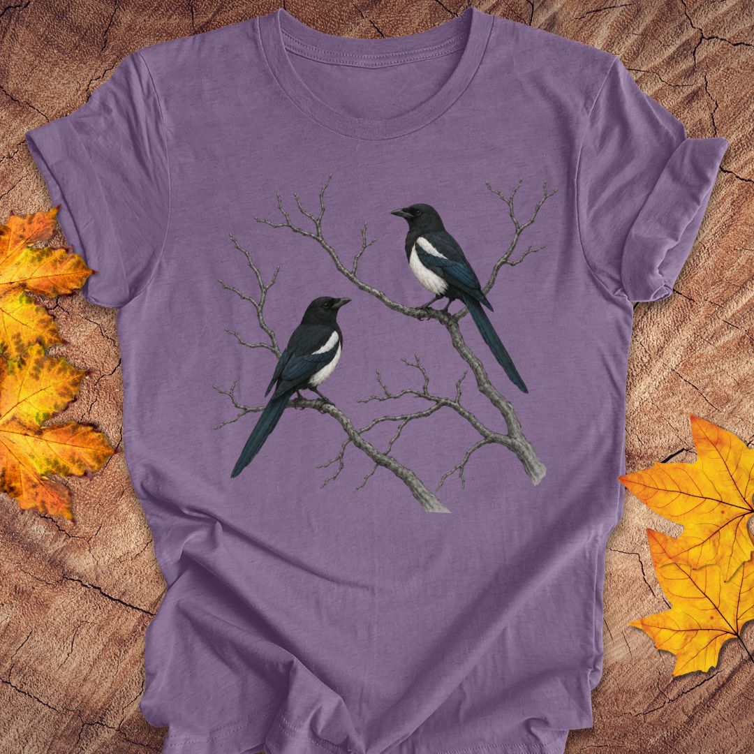 A purple short-sleeve t-shirt with a graphic of two magpies on bare branches, laid flat on a rustic wood background with autumn leaves.