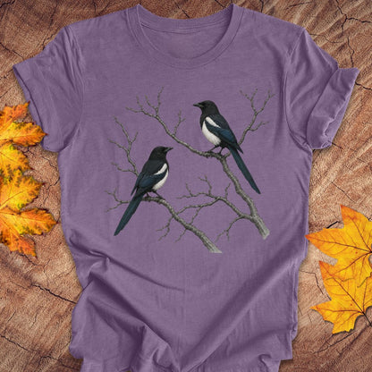 A purple short-sleeve t-shirt with a graphic of two magpies on bare branches, laid flat on a rustic wood background with autumn leaves.