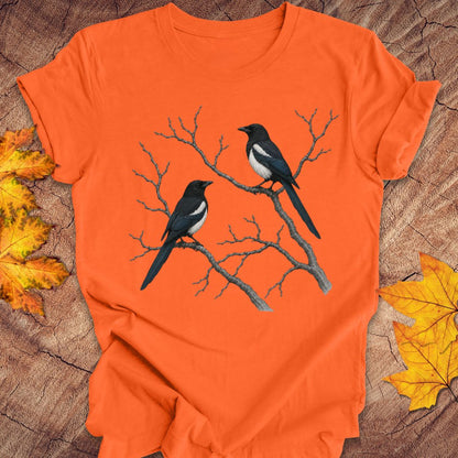 An orange short-sleeve crewneck t-shirt featuring a graphic of two magpies perched on bare tree branches, laid flat on a rustic wood surface with yellow autumn leaves.