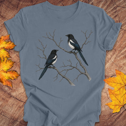 A slate blue crewneck t-shirt featuring a graphic print of two magpies perched on bare tree branches. The shirt is styled in a flat-lay on a rustic wood background with scattered autumn maple leaves.