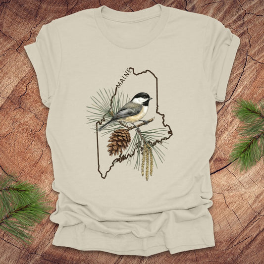 Beige t-shirt with a chickadee and pine cone design inside the outline of the state of maine on a wooden surface