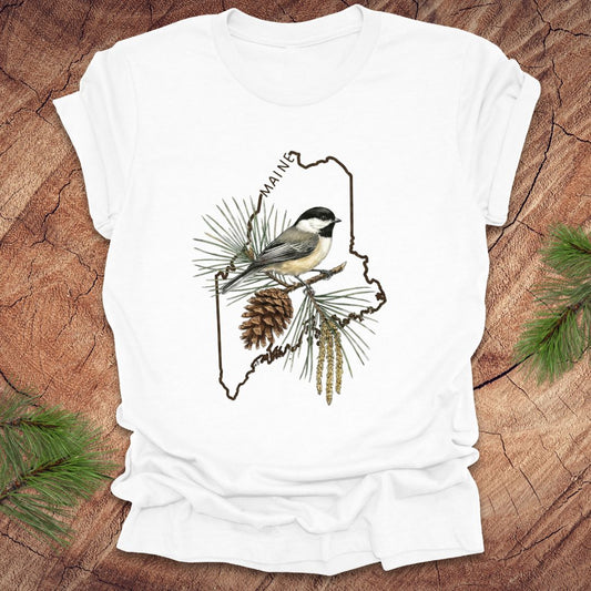 White t-shirt with a chickadee and pine cone design inside the outline of the state of maine on a wooden surface
