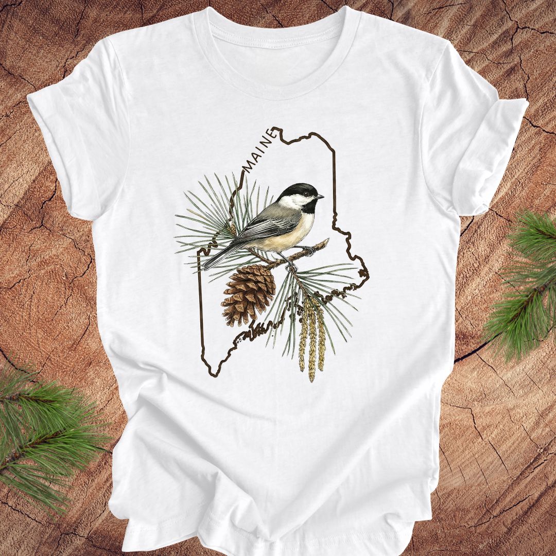 White t-shirt with a chickadee and maine state outline design on a wooden surface