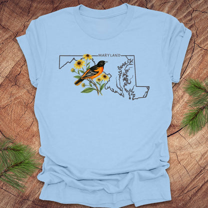 Baby blue t-shirt with Maryland state outline featuring a baltimore oriole and black eyed susans on a wooden surface