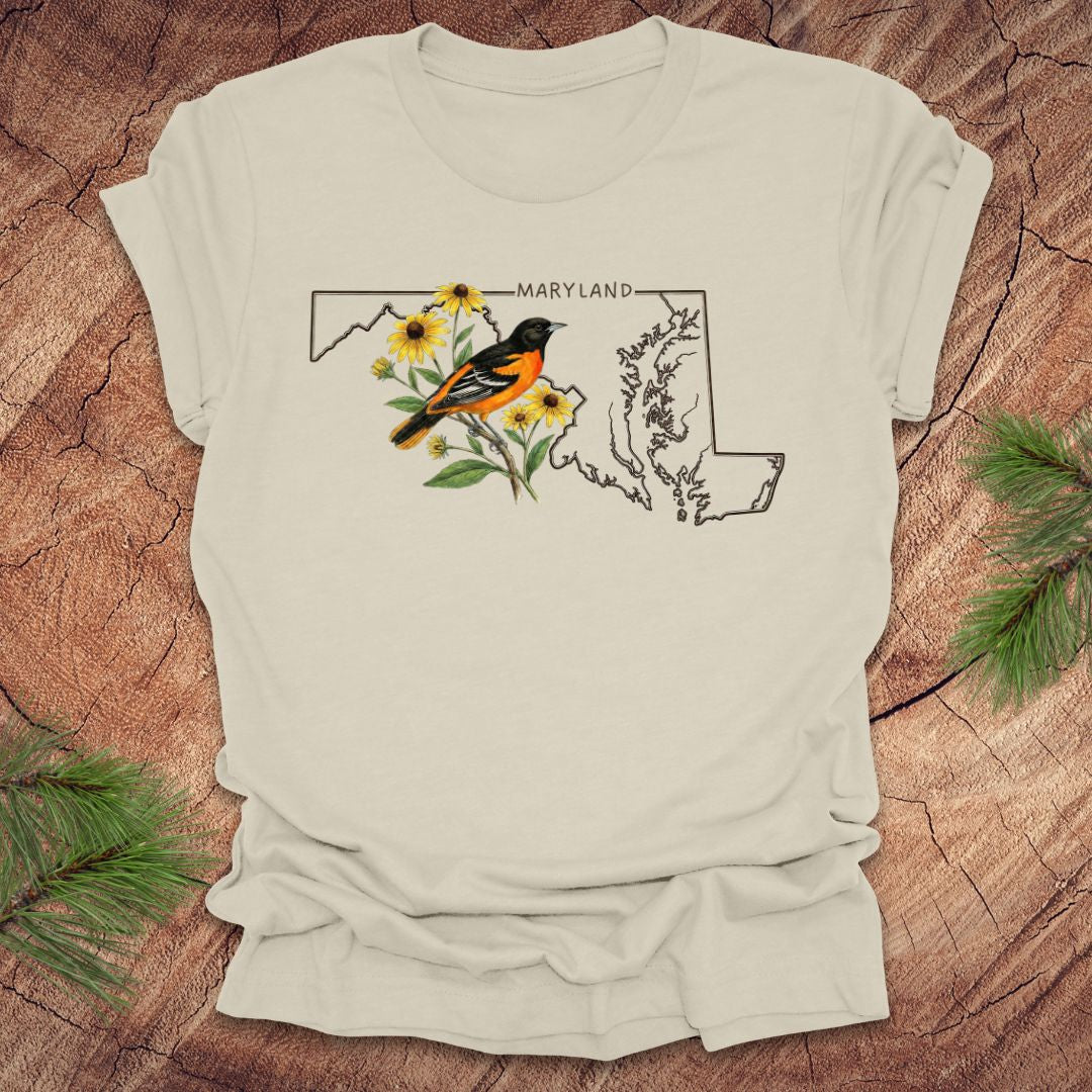 Beige t-shirt with Maryland state outline featuring a baltimore oriole and black eyed susans on a wooden surface