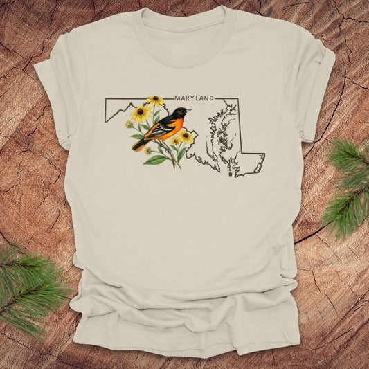 Beige t-shirt with Maryland state outline featuring a baltimore oriole and black eyed susans on a wooden surface