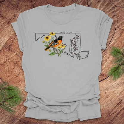 Silver t-shirt with Maryland state outline featuring a baltimore oriole and black eyed susans on a wooden surface