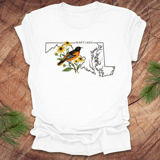 White t-shirt with Maryland state outline featuring a baltimore oriole and black eyed susans on a wooden surface