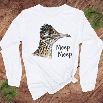 A white long-sleeve t-shirt with a graphic of a roadrunner bird and the text 'Meep Meep' printed on the front, styled flat on a rustic wood background with pine branches.